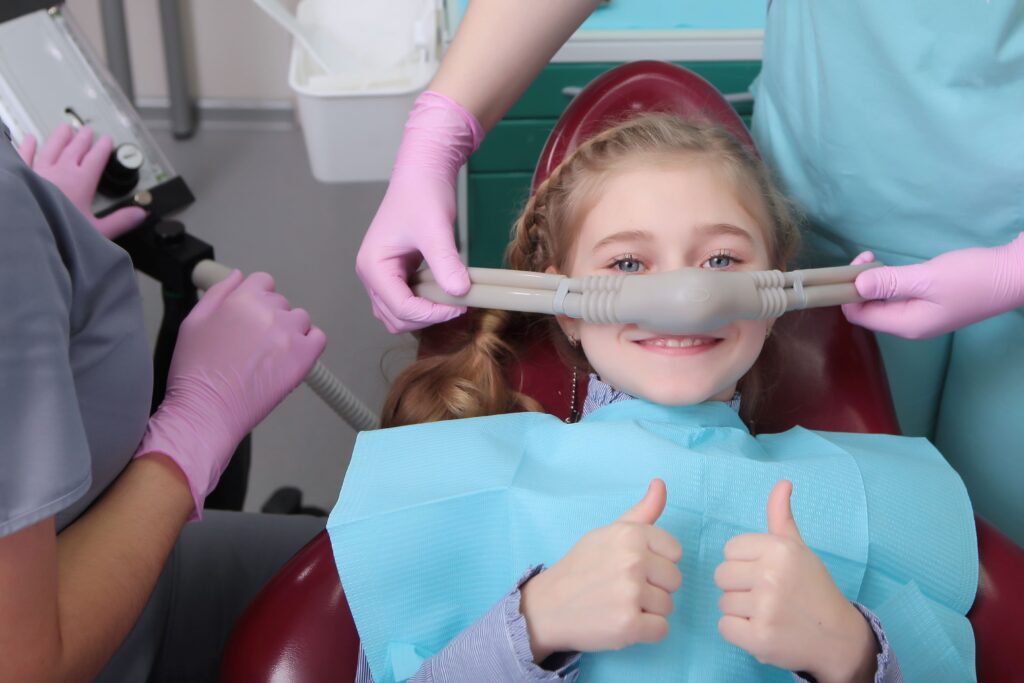 young girl in dentist chair wearing a sedation mask and giving thumbs up